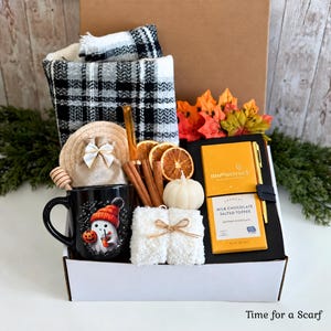 May include: A gift box containing a black and white plaid scarf, a black mug with a ghost graphic, a small burlap bag, cinnamon sticks, dried orange slices, a pumpkin-shaped candle, a white fuzzy item, and a Moonstruck chocolate bar.