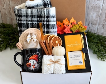 May include: A gift box containing a black and white plaid scarf, a black mug with a ghost graphic, a small burlap bag, cinnamon sticks, dried orange slices, a pumpkin-shaped candle, a white fuzzy item, and a Moonstruck chocolate bar.