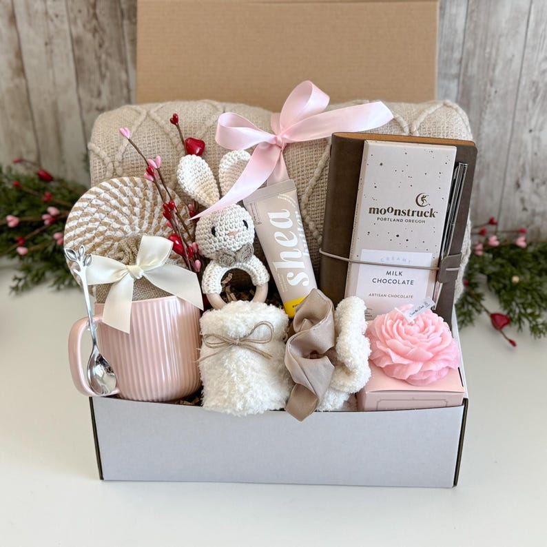 May include: A gift box containing a cream-colored knit blanket, a pink ribbed mug with a silver spoon, a crochet bunny rattle, a tube of shea cream, a brown notebook with a Moonstruck milk chocolate bar, a white fuzzy pouch, and a pink rose candle.