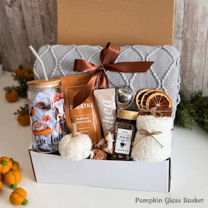 May include: A gift basket containing a gray textured throw blanket with a brown ribbon, a glass tumbler with a bamboo lid and metal straw, a bag of mineral bath soak, a shea cream tube, a heart-shaped tea infuser, herbal tea, and dried orange slices.