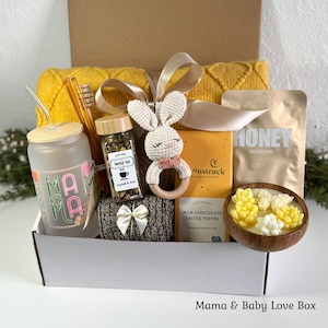 May include: A gift box containing a frosted glass cup with a straw and Mama text, a jar of herbal tea, a crocheted bunny rattle, a bar of Moonstruck milk chocolate, a honey pouch, a wooden bowl with succulent-shaped candles, and a yellow blanket.