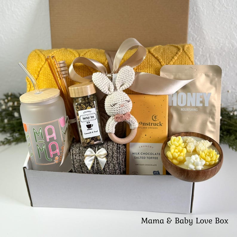 May include: A gift box containing a frosted glass cup with a straw and Mama text, a jar of herbal tea, a crocheted bunny rattle, a bar of Moonstruck milk chocolate, a honey pouch, a wooden bowl with succulent-shaped candles, and a yellow blanket.