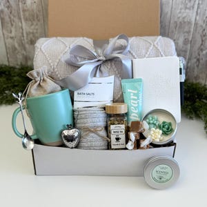 May include: A gift box containing a light gray textured throw blanket with a silver ribbon, a mint green mug, a heart-shaped tea infuser, a silver spoon, bath salts, a white notebook with a tree design, herbal tea, a tube of cream, and a candle.
