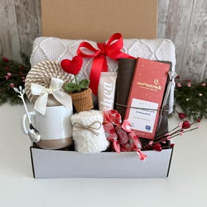 May include: A gift box containing a white and tan mug with a cream bow, a crocheted red heart plant, a white fuzzy towel, a tube of shea cream, a brown journal with a silver pen, a Moonstruck chocolate bar, and heart-shaped candies, tied with a red ribbon.