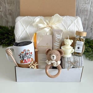 May include: A gift box containing a white mug with a floral MAMA print, a wooden honey dipper, a Moonstruck milk chocolate bar, a shea butter tube, a crochet bear rattle, a bear-shaped candle, herbal tea, and a white textured throw blanket.