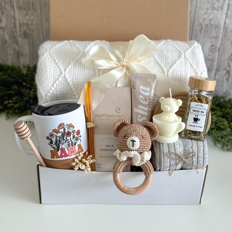 May include: A gift box containing a white mug with a floral MAMA print, a wooden honey dipper, a Moonstruck milk chocolate bar, a shea butter tube, a crochet bear rattle, a bear-shaped candle, herbal tea, and a white textured throw blanket.