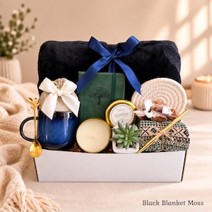 May include: A gift box containing a black plush blanket tied with a blue ribbon, a dark blue ceramic mug with a gold spoon, a green notebook with a tree design, a scented candle in a gold tin, a small succulent, a woven coaster, and a patterned cloth.