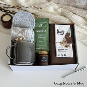May include: A gift box containing a grey ceramic mug, a grey coiled coaster, grey ribbed socks, a green packet of Muscle mineral bath soak, a small brown candle, a brown leather-bound journal, and a Theo organic hazelnut crisp milk chocolate bar.