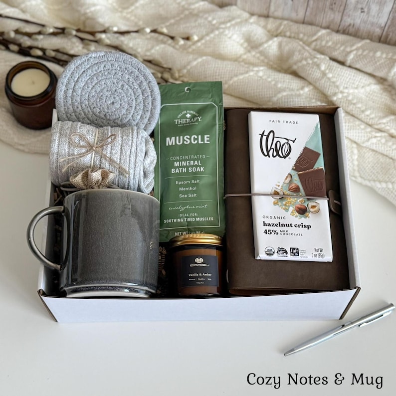 May include: A gift box containing a grey ceramic mug, a grey coiled coaster, grey ribbed socks, a green packet of Muscle mineral bath soak, a small brown candle, a brown leather-bound journal, and a Theo organic hazelnut crisp milk chocolate bar.