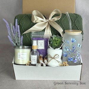 May include: A gift box containing a green knit blanket with a beige ribbon, a glass tumbler with a blue floral print and straw, a candle, a spray bottle, purple bath salts, a small succulent in a white pot, and a white towel tied with twine.