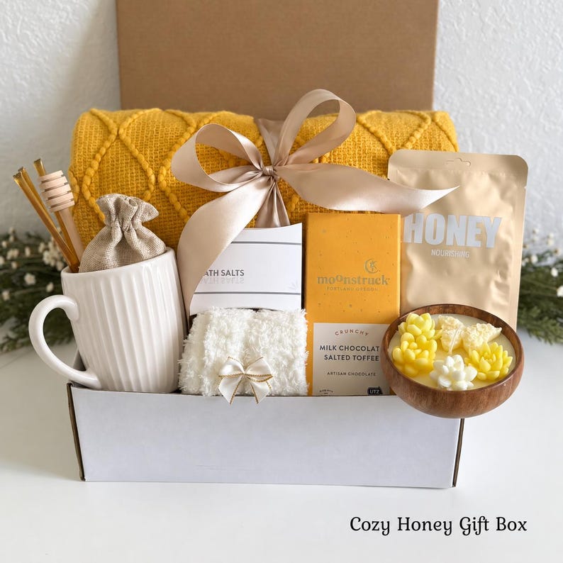 Pu&ograve; includere: Una confezione regalo contenente una coperta gialla testurizzata, una tazza bianca a coste con un bastoncino per miele in legno, un sacchetto beige, sali da bagno, una tavoletta di cioccolato al latte Moonstruck, una bustina di miele e una ciotola in legno.