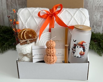 May include: A gift box containing a white textured blanket tied with an orange ribbon, a frosted glass tumbler with a ghost graphic, a stack of three orange pumpkin candles, a Moonstruck milk chocolate bar, bath salts, and dried orange slices.