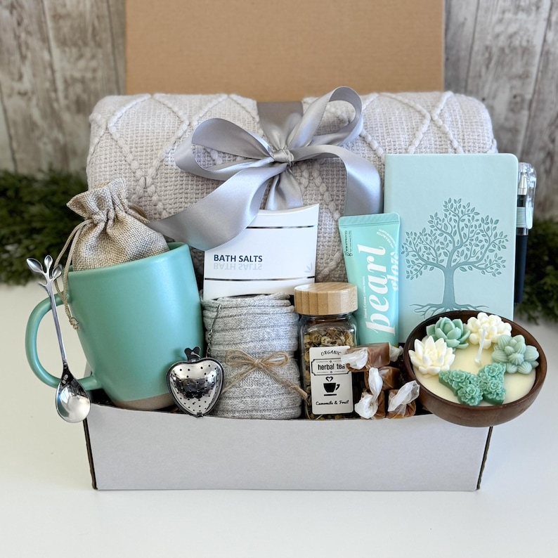 May include: A gift box containing a light green mug, a silver heart-shaped tea infuser, a silver spoon, a textured throw blanket with a grey ribbon, a notebook with a tree graphic, a candle with succulent shapes, herbal tea, and bath salts.