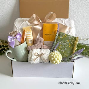 May include: A gift box containing a light green mug with a purple ribbon-tied burlap pouch, a yellow Moonstruck chocolate bar, a green floral notebook, a white bubble candle, fuzzy white socks, and a brown hair clip, all topped with a beige bow.