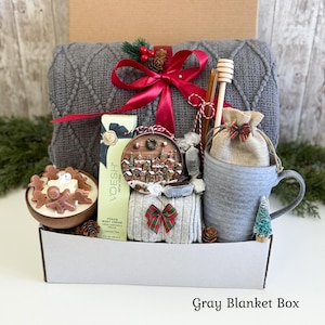 May include: A gift box containing a gray cable-knit blanket tied with a red ribbon, a V'OESH green tea body cream, a round ornament with a winter village scene, a gray mug with a burlap pouch, a wooden honey dipper, and a gingerbread candle.