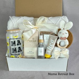 May include: A gift box containing a frosted glass tumbler with sunflower graphics and the text MAMA, a Moonstruck milk chocolate bar, a spray bottle, a tube of shea butter, a bag of bath salts, a crocheted bunny rattle, and a white knit blanket.