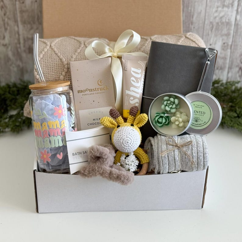 K&ouml;nnte beinhalten: Eine Geschenkbox mit einem Glastrinkbecher mit Bambusdeckel und Strohhalm, einer Moonstruck-Milchschokoladentafel, einer Sheacreme-Tube, einem grauen Notizbuch, einer sukkulentenf&ouml;rmigen Kerze, einer geh&auml;kelten Giraffenrassel, einer flauschigen Haarspange und einem grauen Rippstoff.