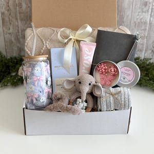 K&ouml;nnte beinhalten: Eine Geschenkbox mit einem Glastrinkbecher mit Schleifenmuster und Bambusdeckel, einer Moonstruck Zartbitterschokolade, einer Rosenhandcreme, einem grauen Notizbuch, einer Kerze mit Blumenwachsverzierungen, einer geh&auml;kelten Elefantenrassel und Haarspange.