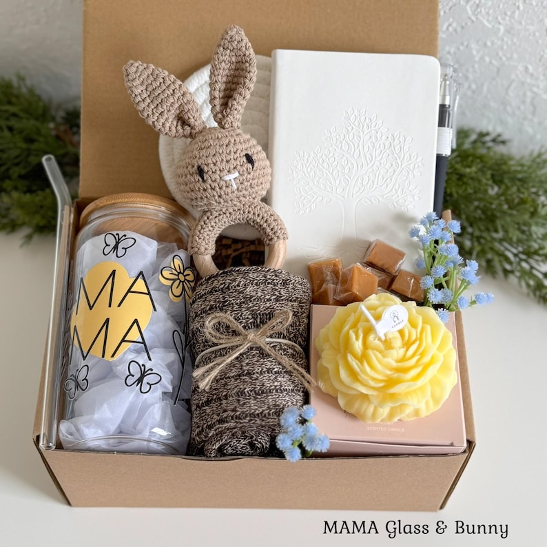 May include: A gift box containing a glass tumbler with a bamboo lid and the text MAMA, a brown crocheted bunny rattle, a white notebook with a tree design, a black pen, a brown knit fabric, wrapped caramels, and a yellow flower-shaped candle.