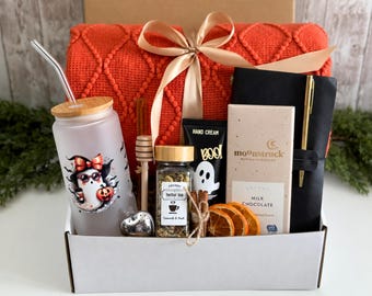 May include: A gift box containing a frosted glass tumbler with a ghost graphic, a wooden honey dipper, a jar of herbal tea, a tube of hand cream, dried orange slices, a Moonstruck milk chocolate bar, a black notebook with a gold pen, and a red blanket.