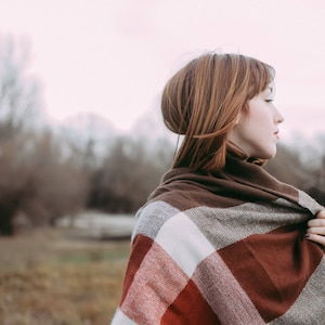 May include: A woman with red hair is wearing a brown, white, and red plaid scarf. The scarf is wrapped around her neck and shoulders. She is standing in a field with trees in the background.