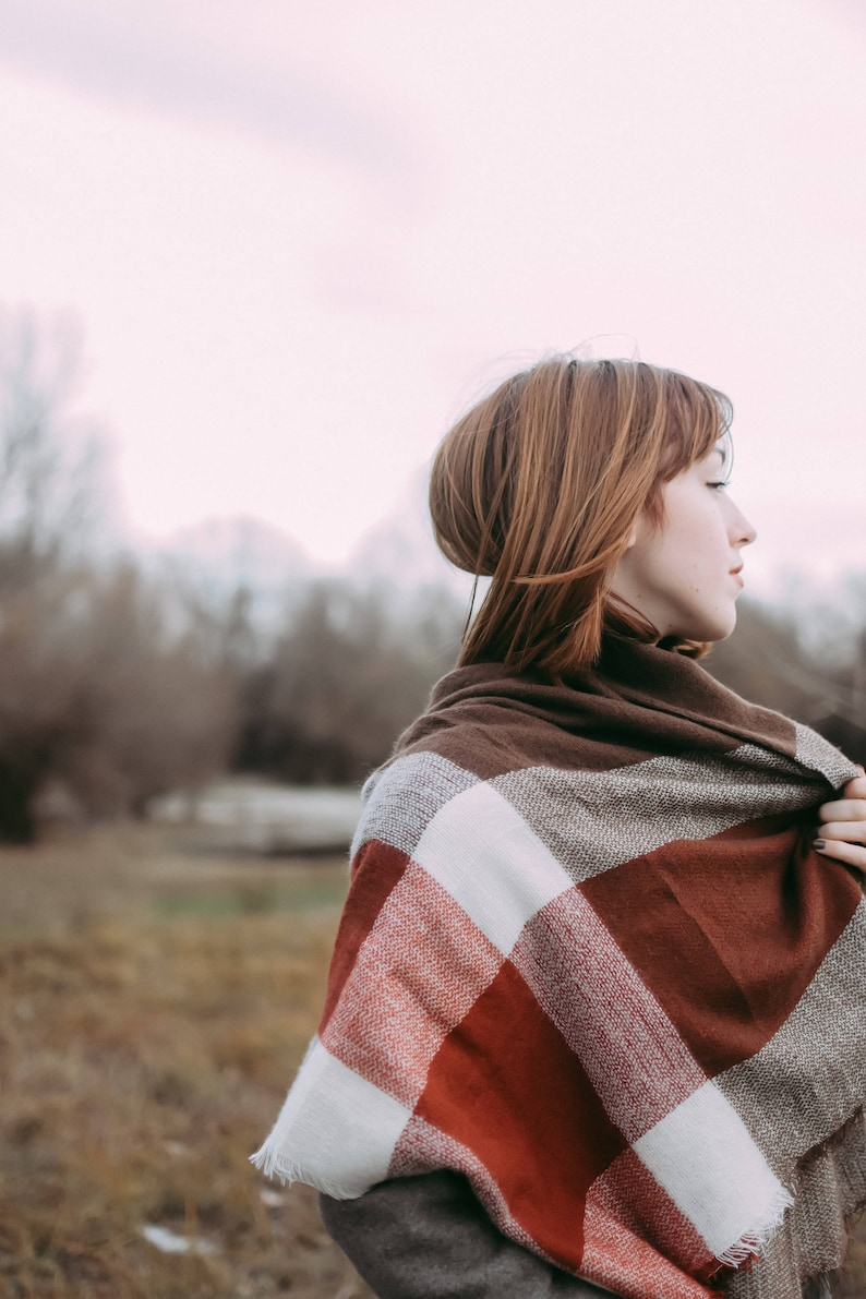 May include: A woman with red hair is wearing a brown, white, and red plaid scarf. The scarf is wrapped around her neck and shoulders. She is standing in a field with trees in the background.