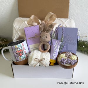 May include: A gift box containing a white mug with a cork base and repeating Mama text, a purple Moonstruck chocolate bar, a crocheted bunny rattle, a white fuzzy cloth, a lavender sage tube, a purple journal, and a wooden bowl with succulent candles.