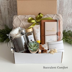 May include: A gift box containing a beige textured throw, a gray ceramic mug with a silver spoon, a heart-shaped tea infuser, a jar of herbal tea, a shea cream tube, a brown notebook with a silver pen, a Moonstruck milk chocolate bar, and bath salts.