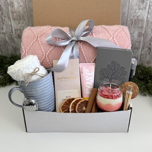 May include: A gift box containing a pink textured throw blanket with a silver ribbon, a grey ribbed mug with a white towel, a Moonstruck milk chocolate bar, a rose hand cream, a grey journal with a tree design, a pen, and a candle with heart wax.