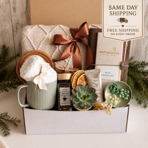 May include: A gift box containing a beige knit blanket with a brown ribbon, a sage green ribbed mug with white fuzzy socks, a jar of herbal tea, dried orange slices, a succulent, a tube of shea cream, a Moonstruck milk chocolate bar, and a candle.