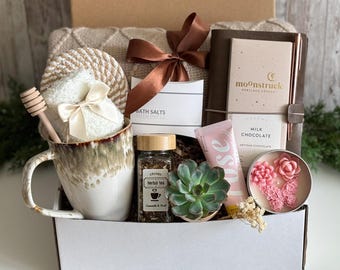 May include: A gift box containing a ceramic mug with a honey dipper, a jar of herbal tea, a succulent, a tube of rose cream, a Moonstruck milk chocolate bar with a pen, bath salts, and a beige knit blanket tied with a brown ribbon.