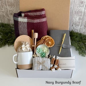 Pu&ograve; includere: Una confezione regalo contenente una sciarpa a quadri bordeaux e bianca, una tazza in ceramica bianca e beige, un taccuino grigio con penna dorata, un bastoncino per miele, fette d'arancia essiccate, cannella, saponi e sali da bagno.