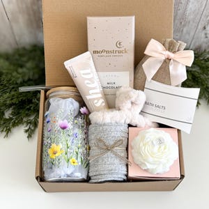May include: A gift box containing a glass tumbler with a floral print, a tube of shea hand cream, a Moonstruck milk chocolate bar, a burlap pouch with a pink ribbon, a white bath salt packet, a fuzzy headband, and a white flower-shaped candle.