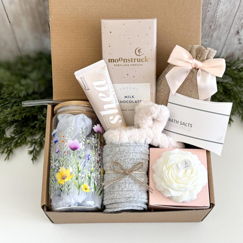 May include: A gift box containing a glass tumbler with a floral print, a tube of shea hand cream, a Moonstruck milk chocolate bar, a burlap pouch with a pink ribbon, a white bath salt packet, a fuzzy headband, and a white flower-shaped candle.