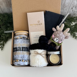 K&ouml;nnte beinhalten: Eine Geschenkbox mit einem Glastrinkbecher mit Bambusdeckel und dem Wort mama in Schwarz, einer Moonstruck-Milchschokolade, einem schwarzen Notizbuch mit silbernem Stift, einem geh&auml;kelten Rentier, flauschigen wei&szlig;en Socken und einer kleinen Kerze.