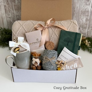 May include: A gift box containing a beige knit blanket with a satin ribbon, a gray ceramic mug with a gold spoon, a Moonstruck chocolate bar, a jar of caramels, a dark green journal with a tree emblem, bath salts, and a shea butter tube.