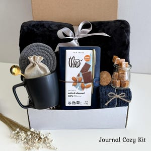 May include: A gift box containing a black plush blanket with a silver ribbon, a dark blue journal with a gold pen, a Theo organic salted almond chocolate bar, a black mug, a gray coaster, a small jar of caramels, and navy blue socks.
