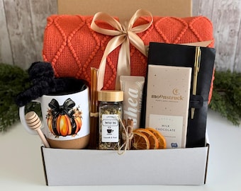 May include: A gift box containing an orange textured blanket with a gold ribbon, a white mug with a pumpkin graphic, a honey dipper, a jar of herbal tea, a tube of shea butter, dried orange slices, a Moonstruck milk chocolate bar, and a black journal with a gold pen.