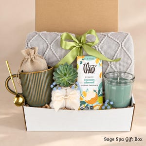 Pu&ograve; includere: Una confezione regalo contenente una coperta grigia testurizzata con un nastro verde, una tazza verde a coste con un cucchiaio dorato, un piccolo sacchetto di juta, una pianta succulenta, una tavoletta di cioccolato Theo e una candela verde.