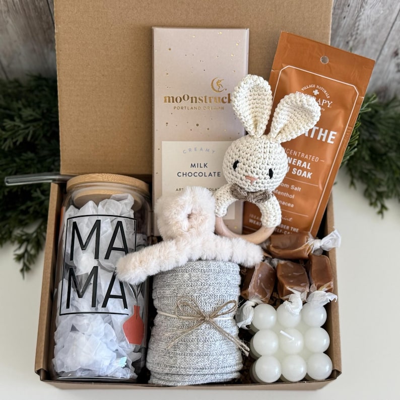 May include: A gift box containing a glass tumbler with a bamboo lid and the text MAMA, a Moonstruck milk chocolate bar, a crocheted bunny rattle, a fuzzy hair claw, a bag of mineral soak, several wrapped caramels, and a white bubble candle.