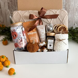 K&ouml;nnte beinhalten: Eine Geschenkbox mit Pflegeprodukten. Sie enth&auml;lt ein Glas mit Halloween-Design, ein Badesalz-P&auml;ckchen, Sheabutter-Lotion, Kr&auml;utertee, einen dekorativen K&uuml;rbis und eine Decke, die mit einem braunen Band gebunden ist. Die Box ist wei&szlig;.