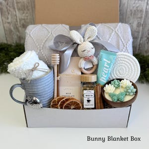 May include: A gift box containing a grey textured blanket with a grey ribbon, a crochet bunny rattle, a grey mug with a heart-shaped tea infuser, a Moonstruck milk chocolate bar, dried orange slices, herbal tea, cinnamon sticks, and a succulent candle.