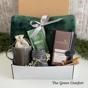 May include: A gift box containing a dark green plush blanket with a silver ribbon, a gray ceramic mug with a small beige drawstring pouch, a packet of Muscle mineral bath soak, a dark gray knit item, a Moonstruck dark chocolate bar, and a honey dipper.