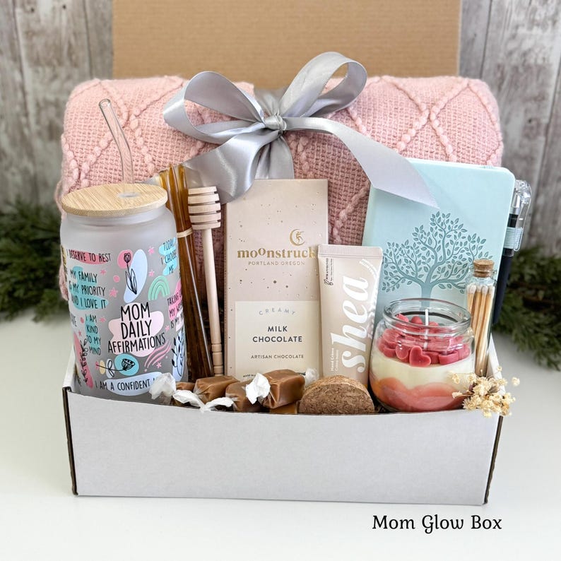 May include: A gift box containing a frosted glass tumbler with Mom Daily Affirmations text, a wooden honey dipper, a Moonstruck milk chocolate bar, a shea cream tube, a light blue journal, a candle with heart wax melts, and a pink knit blanket.