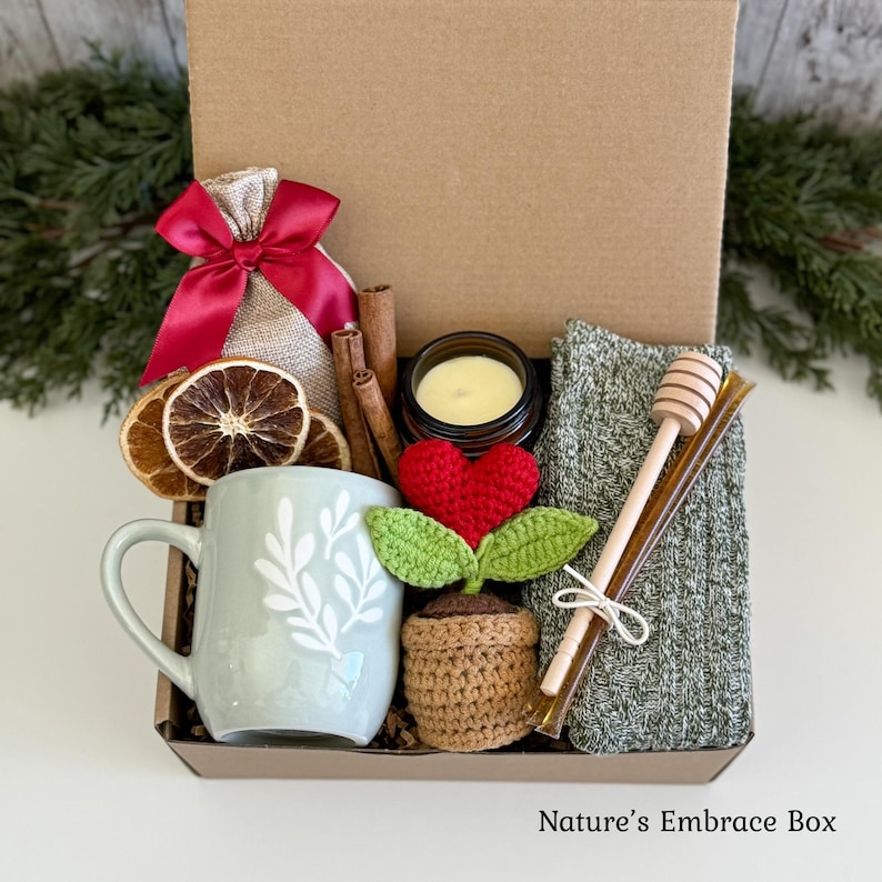 May include: A gift box containing a light green mug with white leaf patterns, a crocheted heart plant in a brown pot, a burlap bag with a red satin bow, dried orange slices, cinnamon sticks, a wooden honey dipper, and a green knit fabric.