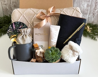May include: A gift box filled with self-care items. Includes a gray mug, a journal with a gold pen, a small succulent, a jar of caramel candies, a shea butter tube, a small blanket, and a decorative coaster. The box is tied with a gold ribbon. Text on the box reads "Care & Cozy Gift Box."