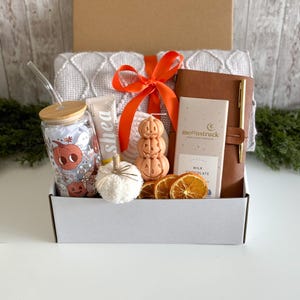 K&ouml;nnte beinhalten: Eine Geschenkbox mit herbstlichen Artikeln. Enth&auml;lt ein Glas mit K&uuml;rbisdesign, Shea-Lotion, einen wei&szlig;en Deko-K&uuml;rbis, eine k&uuml;rbisf&ouml;rmige Kerze, eine Schokoladentafel, ein ledergebundenes Notizbuch und einen Stift. Die Box ist mit einem orangefarbenen Band gebunden.