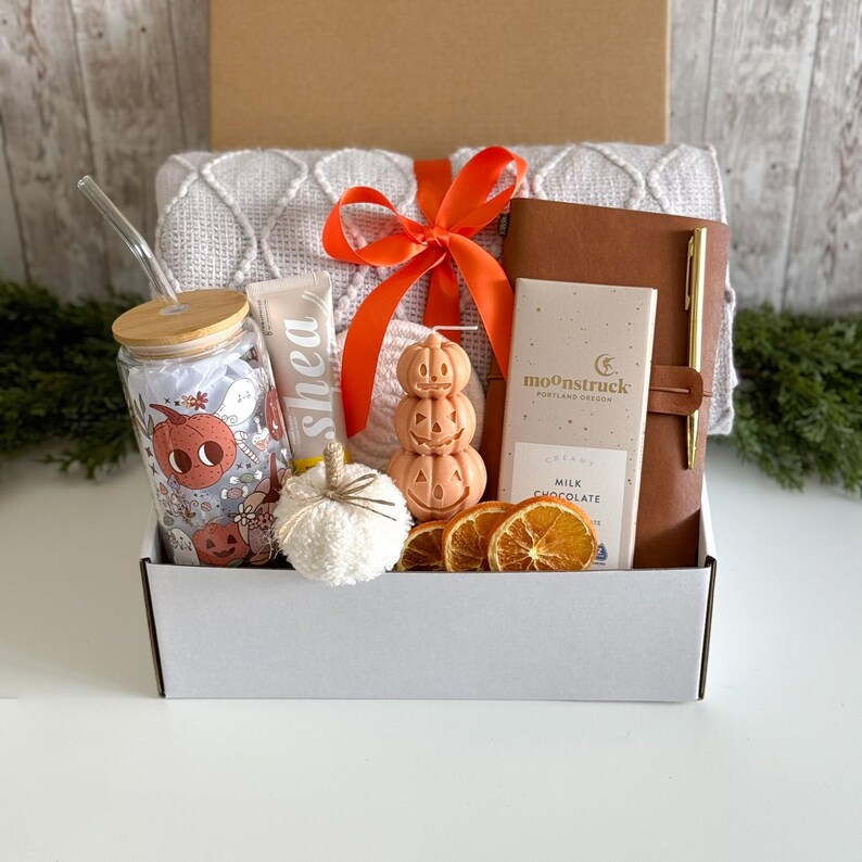 K&ouml;nnte beinhalten: Eine Geschenkbox mit herbstlichen Artikeln. Enth&auml;lt ein Glas mit K&uuml;rbisdesign, Shea-Lotion, einen wei&szlig;en Deko-K&uuml;rbis, eine k&uuml;rbisf&ouml;rmige Kerze, eine Schokoladentafel, ein ledergebundenes Notizbuch und einen Stift. Die Box ist mit einem orangefarbenen Band gebunden.