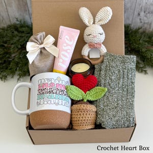 May include: A gift box containing a white mug with a cork base and the repeated text mama, a pink rose hand cream tube, a cream-colored crochet bunny rattle, a small candle, a brown crochet potted heart plant, and a pair of green knit socks.