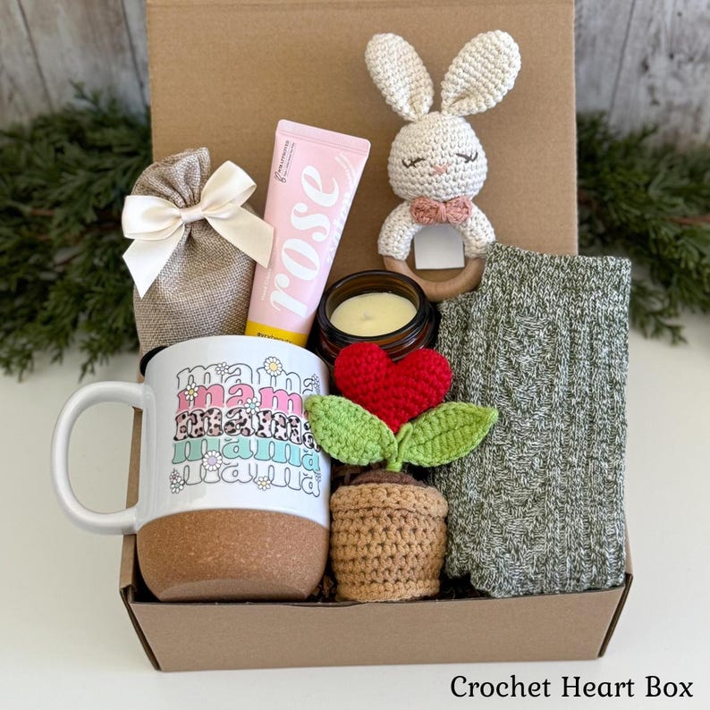 May include: A gift box containing a white mug with a cork base and the repeated text mama, a pink rose hand cream tube, a cream-colored crochet bunny rattle, a small candle, a brown crochet potted heart plant, and a pair of green knit socks.